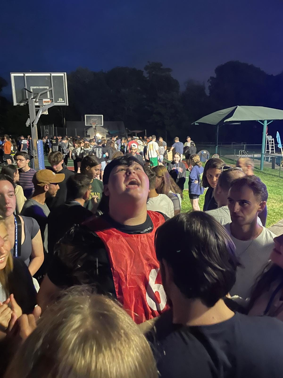 Rafi, foregrounds, gathers with fellow staff members at camp. They stand on a basketball court near green grass, It is evening and the sky is dark but faces are lit with electric lights.