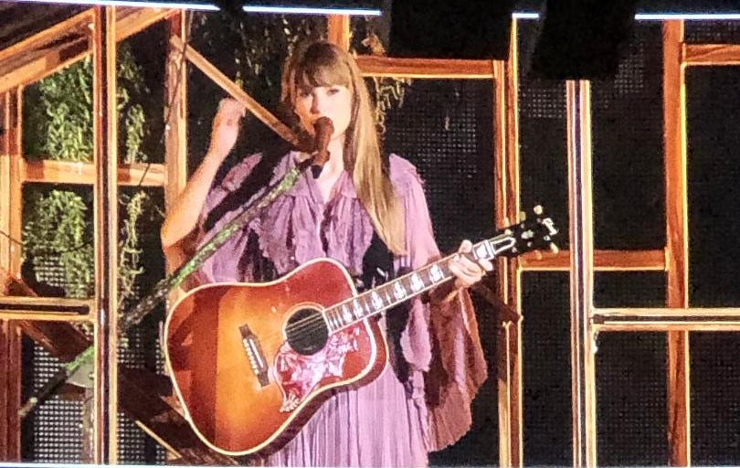 T Swift with acoustic guitar and "Folklore cabin" set, flowing purple dress