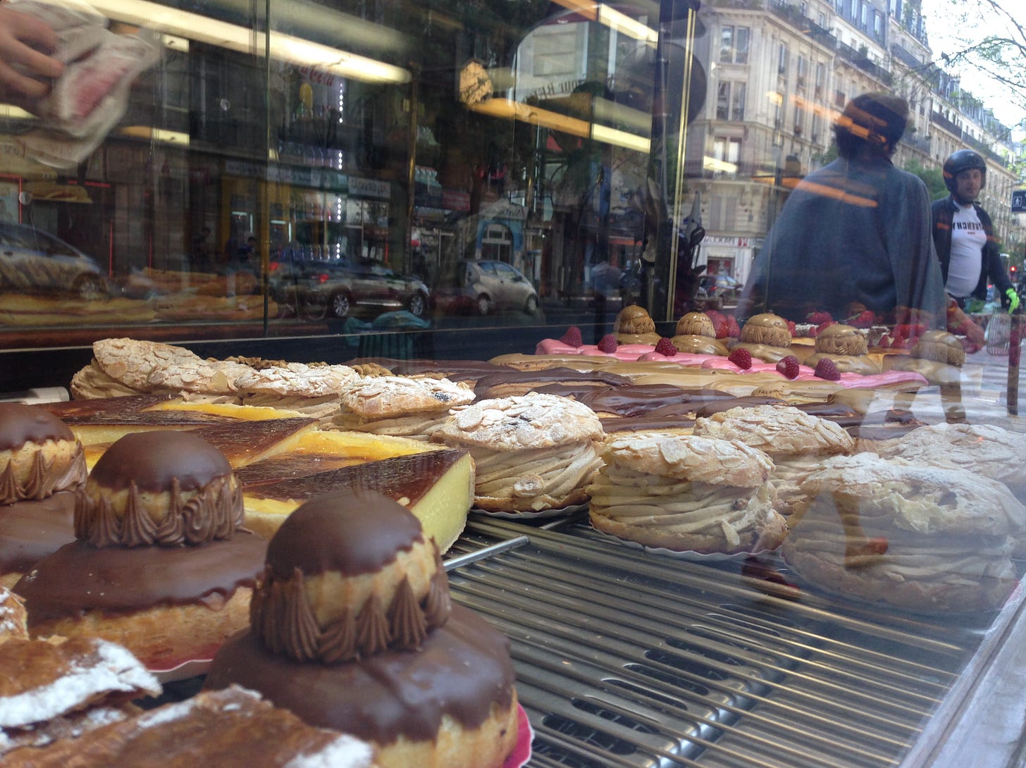 A imagem mostra a vitrine de uma padaria francesa repleta de doces da confeitaria local. No vidro é possível ver o reflexo de pedestres.
