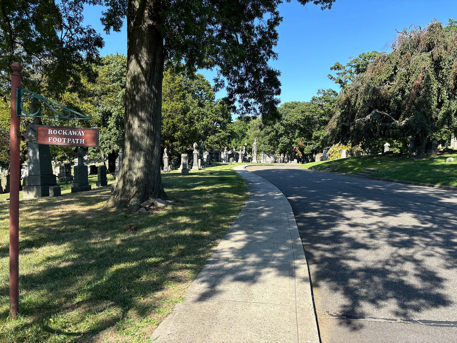 A road in the Evergreens Cemetery. A decorative wood sign suspended from a green metal post reads ROCKAWAY FOOTPATH.