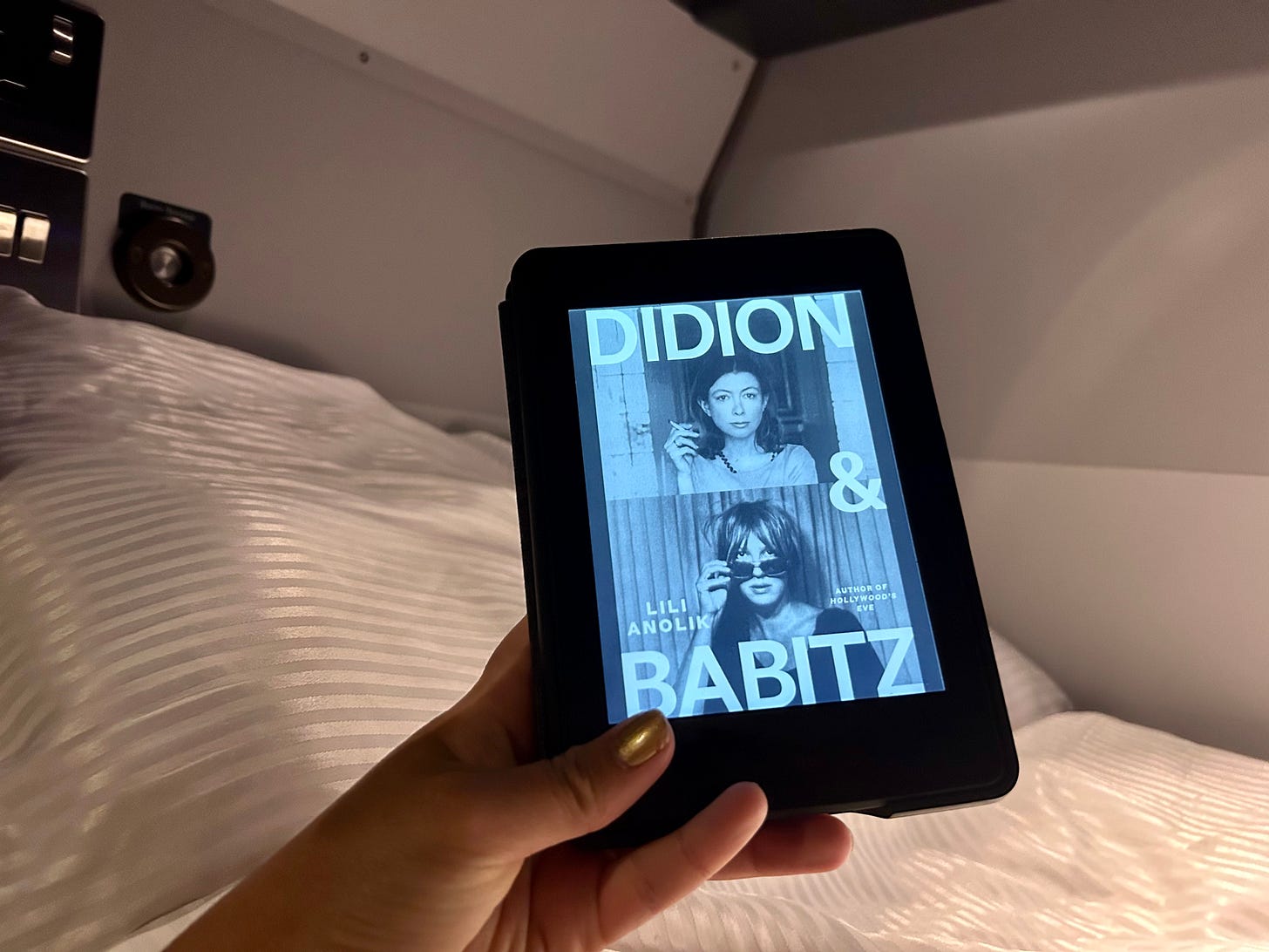 A hand holding a Kindle, featuring the cover of ‘Didion & Babitz’ by Lili Anolik, in front of a bed.