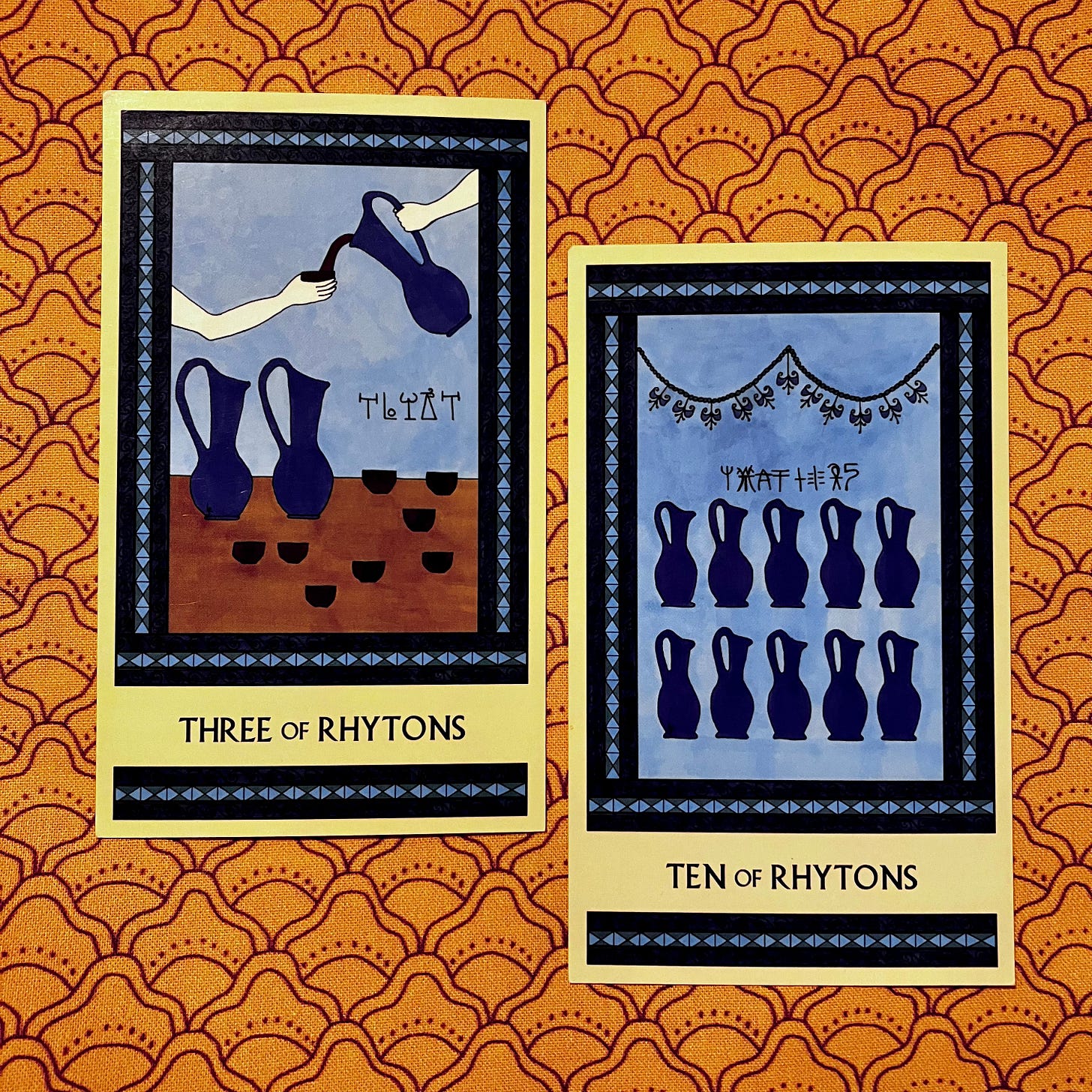 Two Minoan Tarot cards side by side on a gold print fabric background. Both cards are in shades of blue. The Ten of Rhytons shows ten blue pitchers set out in rows beneath a garland of lilies. The Three of Rhytons shows one hand holding a cup while another person's hand fills that cup from a pitcher. More cups and pitchers await nearby.