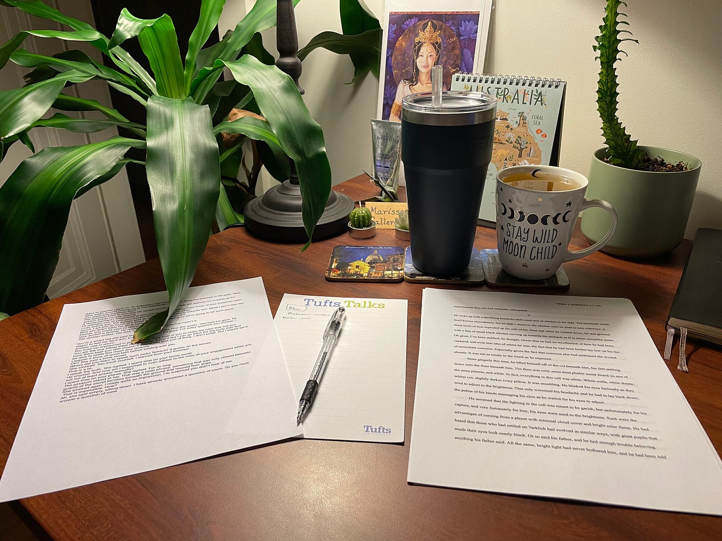 A dimly lit desk with a glass of water and a mug of tea on it shows multiple printed pages with words on them and a notepad and pen. There are plants in the background. 