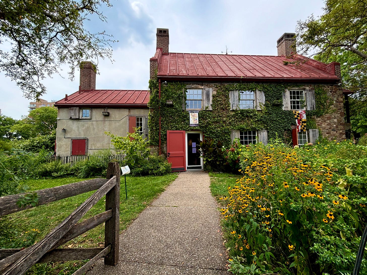 A Colonial Dutch-looking house. The front is covered in ivy. The garden is blooming in such abundance you can almost hear the hum of the bees. Outside the front window, the Maryland state flat is hung against the ivy-covered wall. An apartment building is just visible behind the trees to the left of the house.