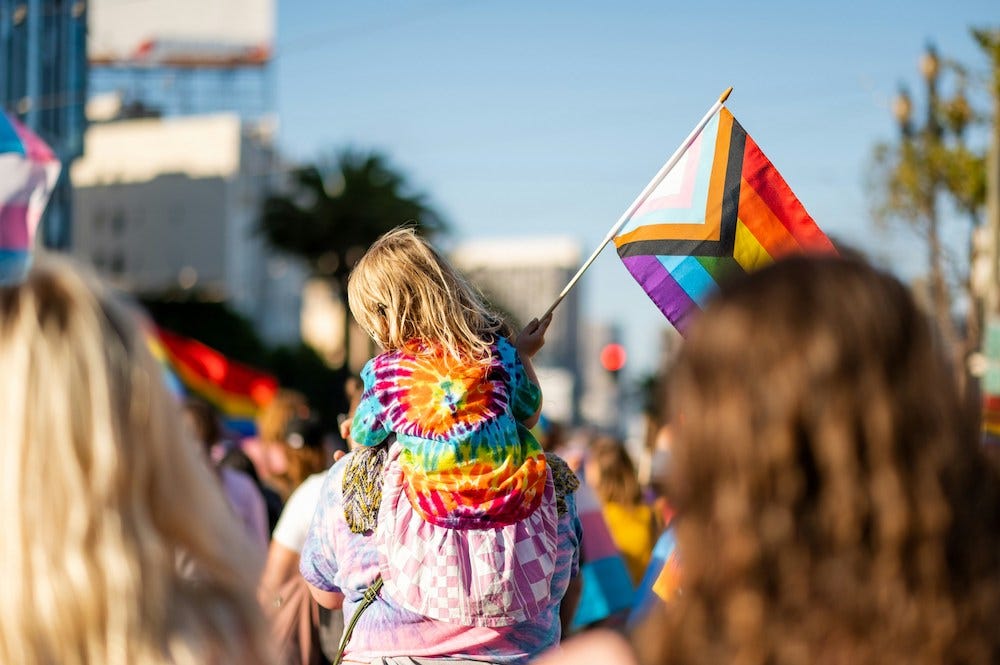 A pride march featuring a child carrying a progress pride flag riding on the shoulders of an adult on a summer’s day