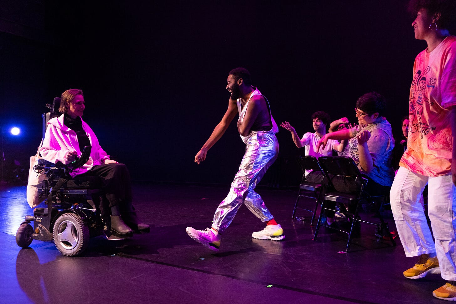 Jerron dances with Park McArthur, a White wheelchair user, as Carolyn Lazard, a Brown nonbinary person, stands behind. Seated audience bops along.