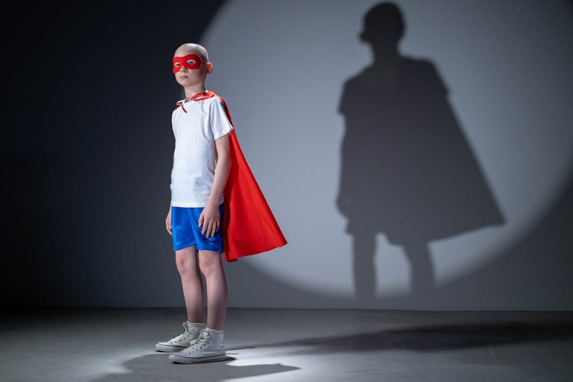 Free Child in a superhero costume poses confidently with a shadow on a studio wall, symbolizing strength and dreams. Stock Photo