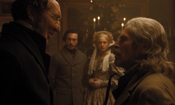Two older men with glasses discuss intently while a young couple looks on from the background.