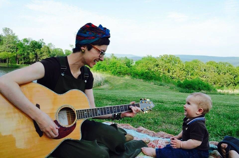 Kaitlin is playing guitar and singing to her son who is about 1 1/2 years old. He is smiling up at her. They are sitting on grass with Ozark mountains in the background