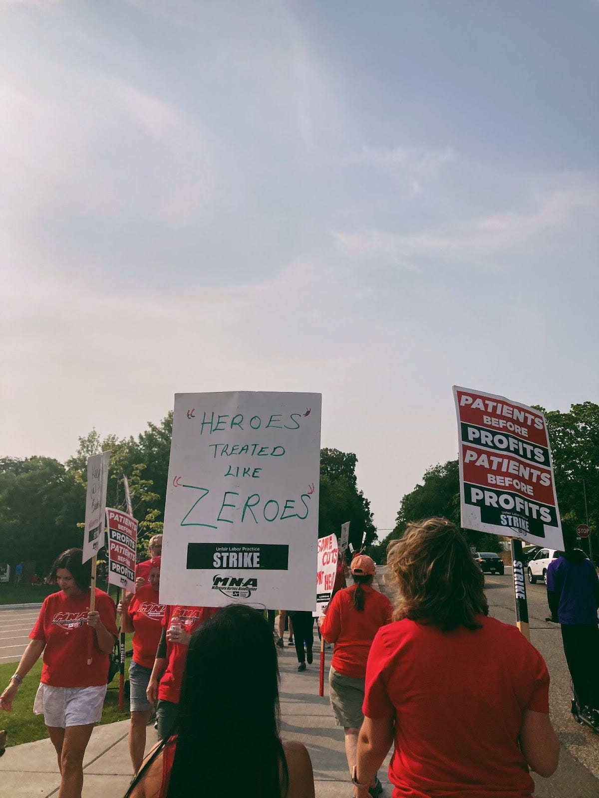 picketers wearing red shirts carry signs that read "heroes treated like zeroes" and "patients before profits"