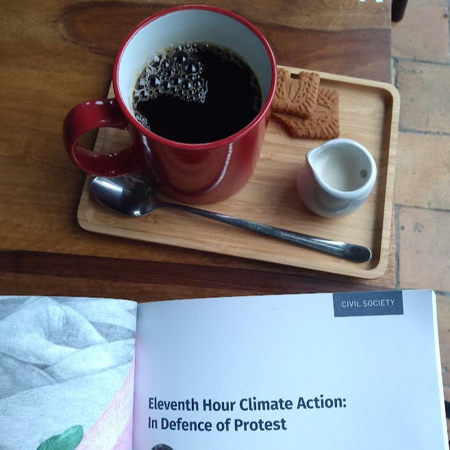 Photo of a mug of coffee on a wooden tray on a table, with the edge of a book open on the title 'In defence of climate protest'