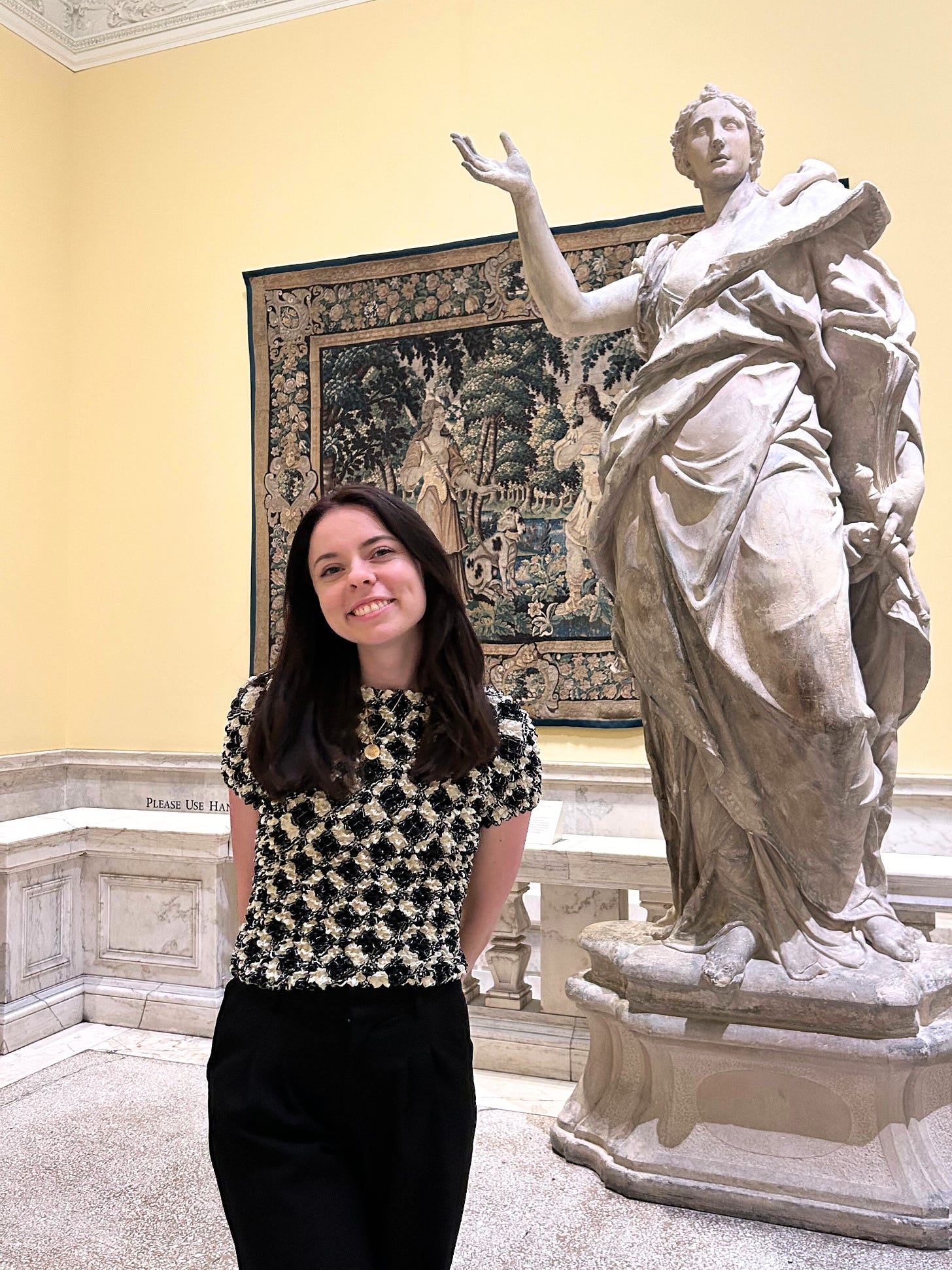 Sierra stands smiling next to a classical statue at the Walters Museum in Baltimore, Maryland, with a decorative tapestry displayed in the background.