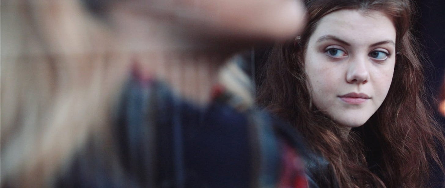 Georgie Henley looks to the left, towards another actress who appears out of focus in the front of the frame.
