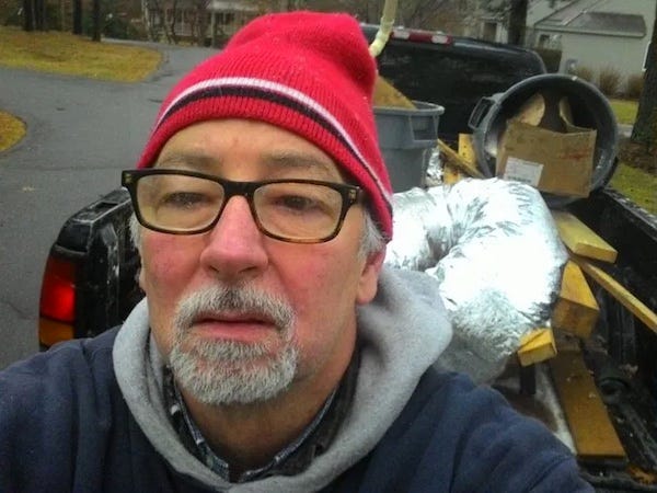 selfie of a sixty year old man, his face brigh red from the cold, wearing work clothes and a ski cap, standing in front of a pickup truck full of demolition debris.
