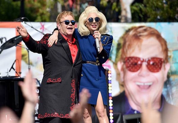 Two performers on stage: one in a decorated suit with sunglasses, the other in a stylish dress with a microphone, both smiling and embracing