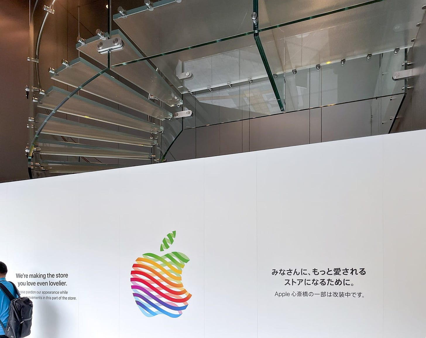 An interior shot of the first floor at Apple Shinsaibashi. A large white wall with the heritage logo blocks access to the second level and spiral glass staircase. The wall says "We're making the store you love even lovelier."