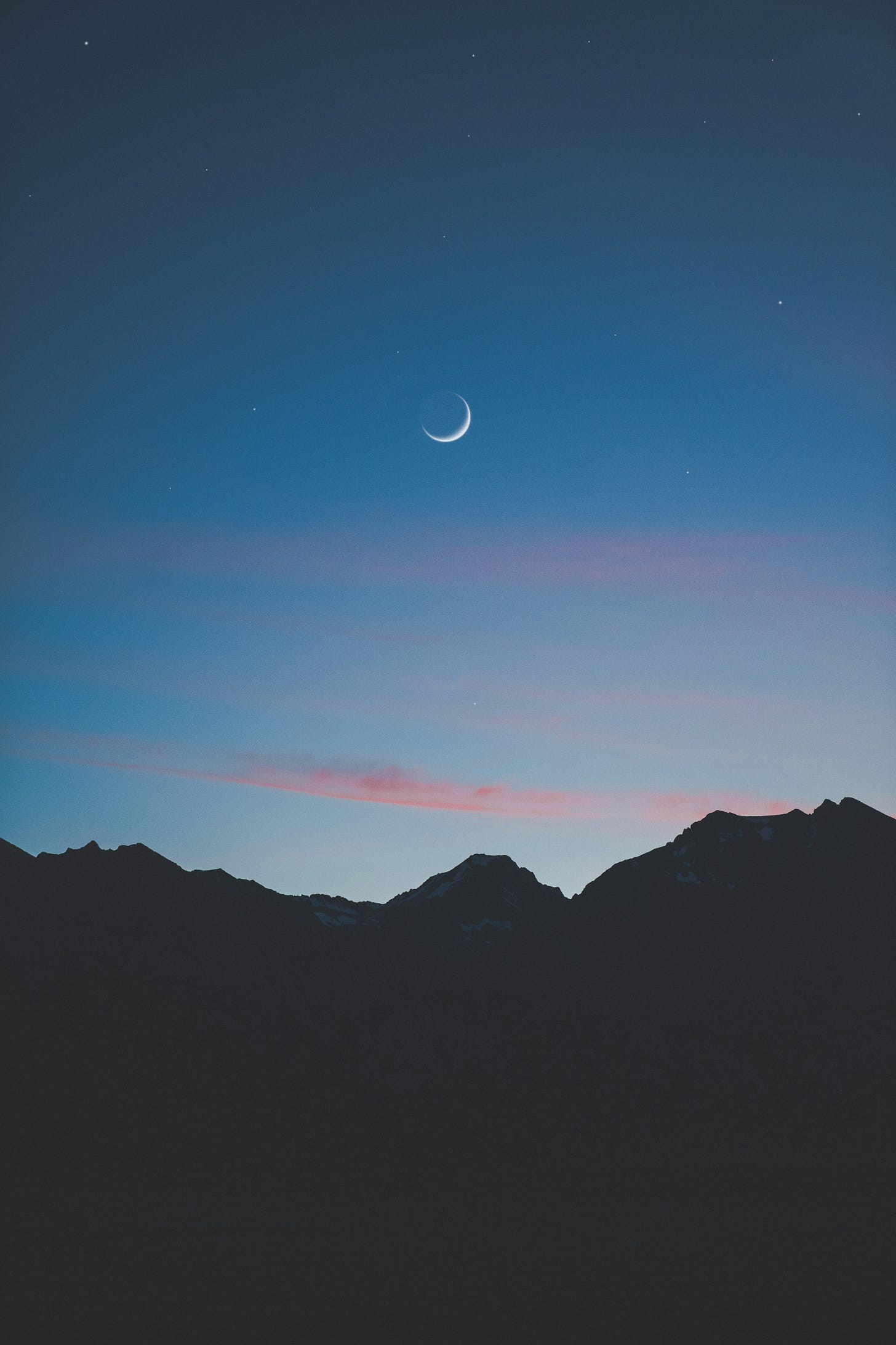 Silhouetted mountain peaks under a darkening sky with pink clouds near the horizon and a crescent moon further up.