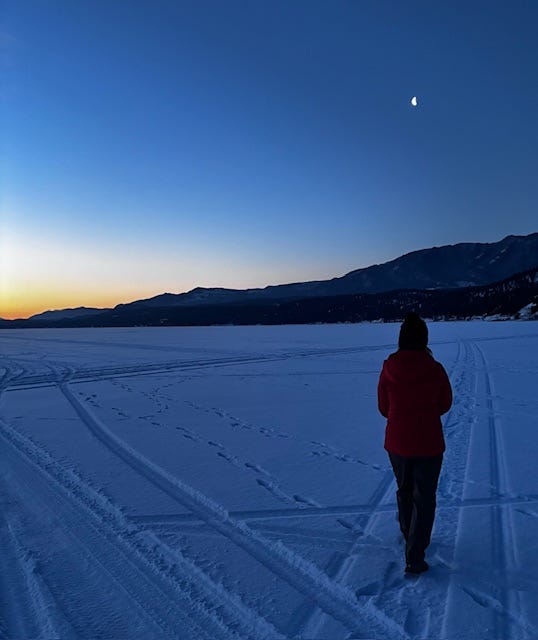 Woman walking on frozen, snowy lake that is surrounded by mountains at sunrise.