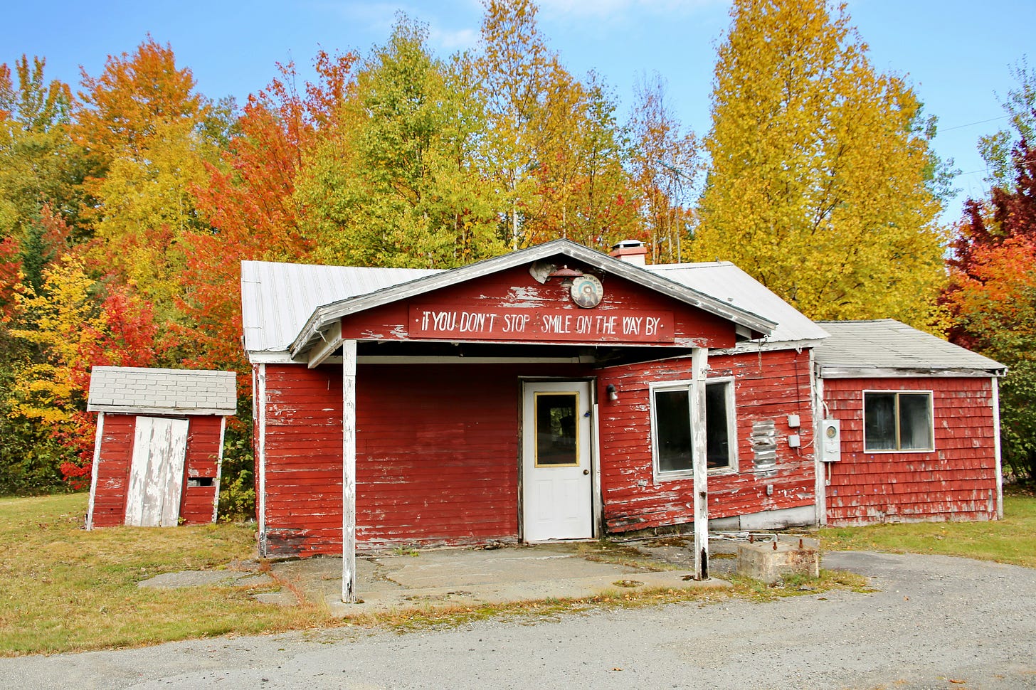 Dilapidated red building and outbuilding in front of trees with red, orange and yellow leavees.