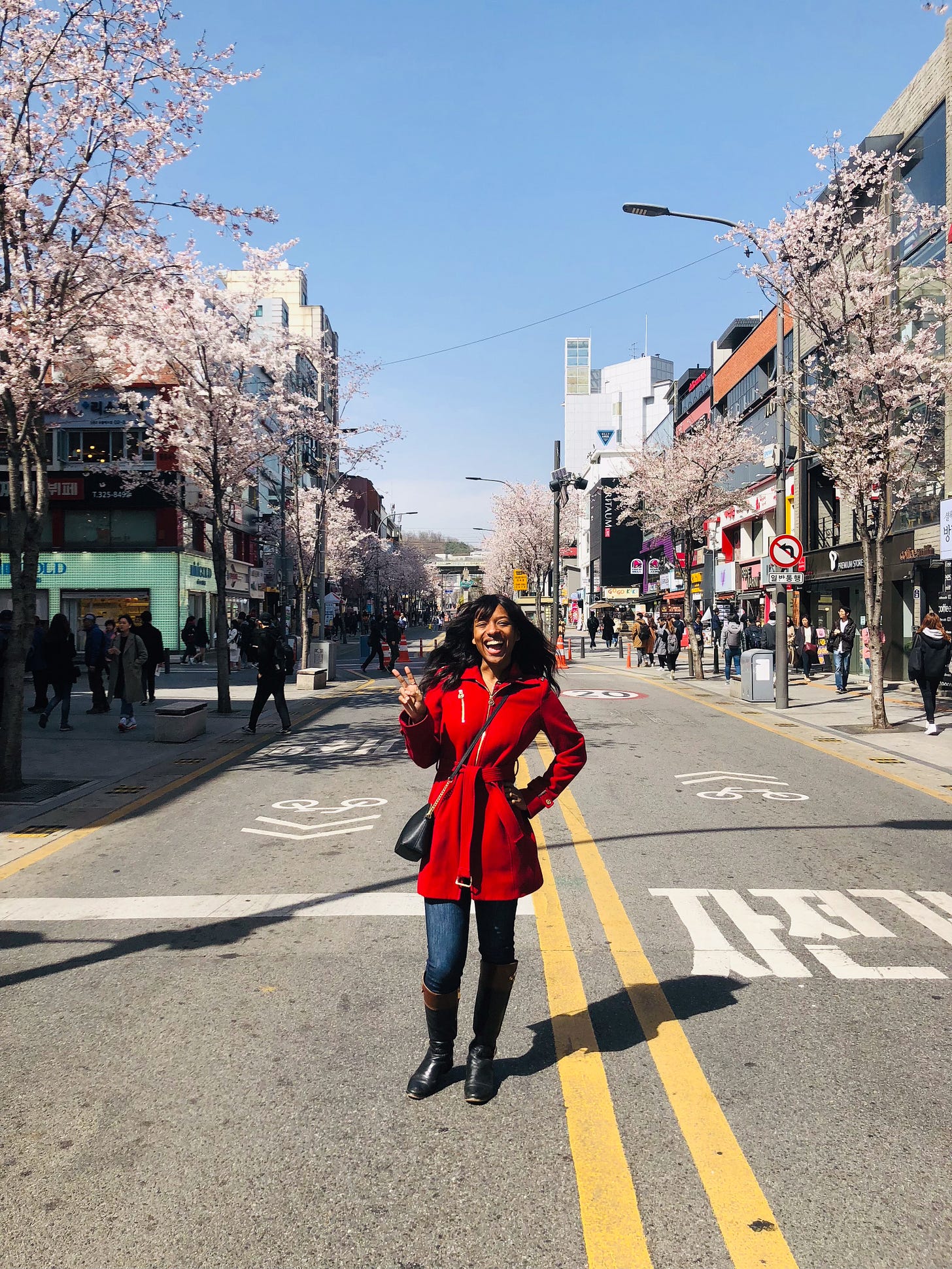 A photo of Kat Lewis, a Black woman wearing a hot-colored trench coat, cool-colored jeans, and tall boots. She stands in the middle of a street in Sinchon, Seoul, South Korea. The street is lined with cherry blossom trees in full bloom.