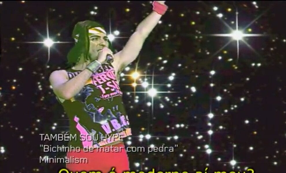 Cena da esquete "Também Sou Hype", que mostra o saudoso humorista Fausto Fanti, do Hermes e Renato, de peruca, vestido com roupas de hipster dos anos 2000. Ele está num fundo de estrelas. O texto na imagem diz: "bichinho de matar com pedra". A estética lembra muito a estética vintage de fitas VHS, quando o tempo degrada levemente a qualidade da imagem.