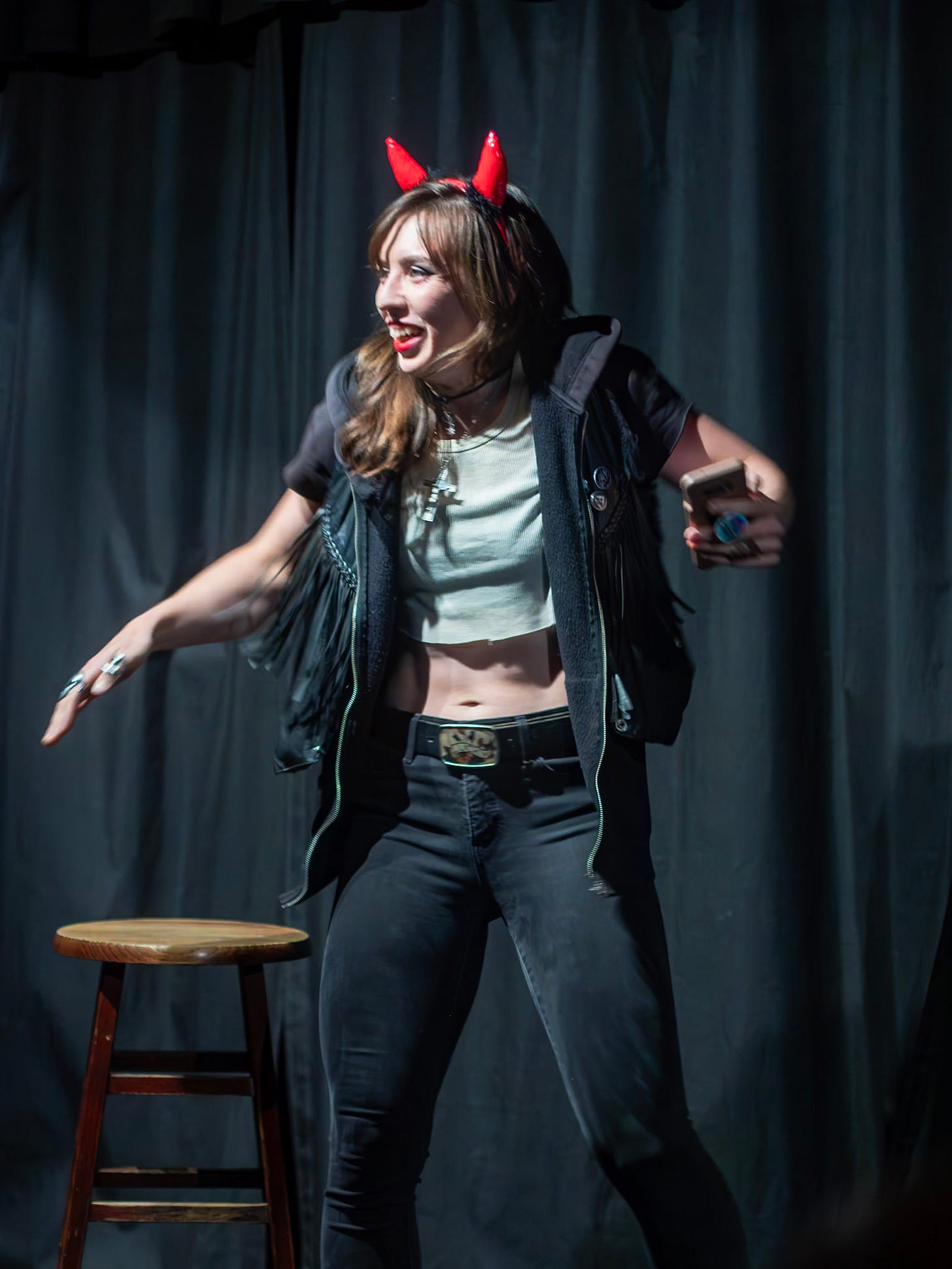 Cara stands on a stage, wearing red devil horns, jeans, and a black leather hoodie and vest, she is gesturing and smiling wildly.