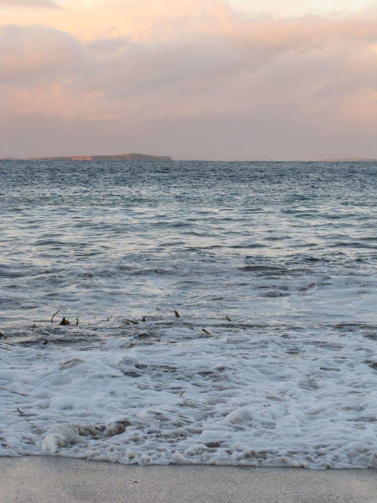 A pastel image of a foamy sea, with seaweed caught in the waves beyond the shore. The sky is tinged with colour by the slow sunlight, and the islands of Orkney lay on the horizon.
