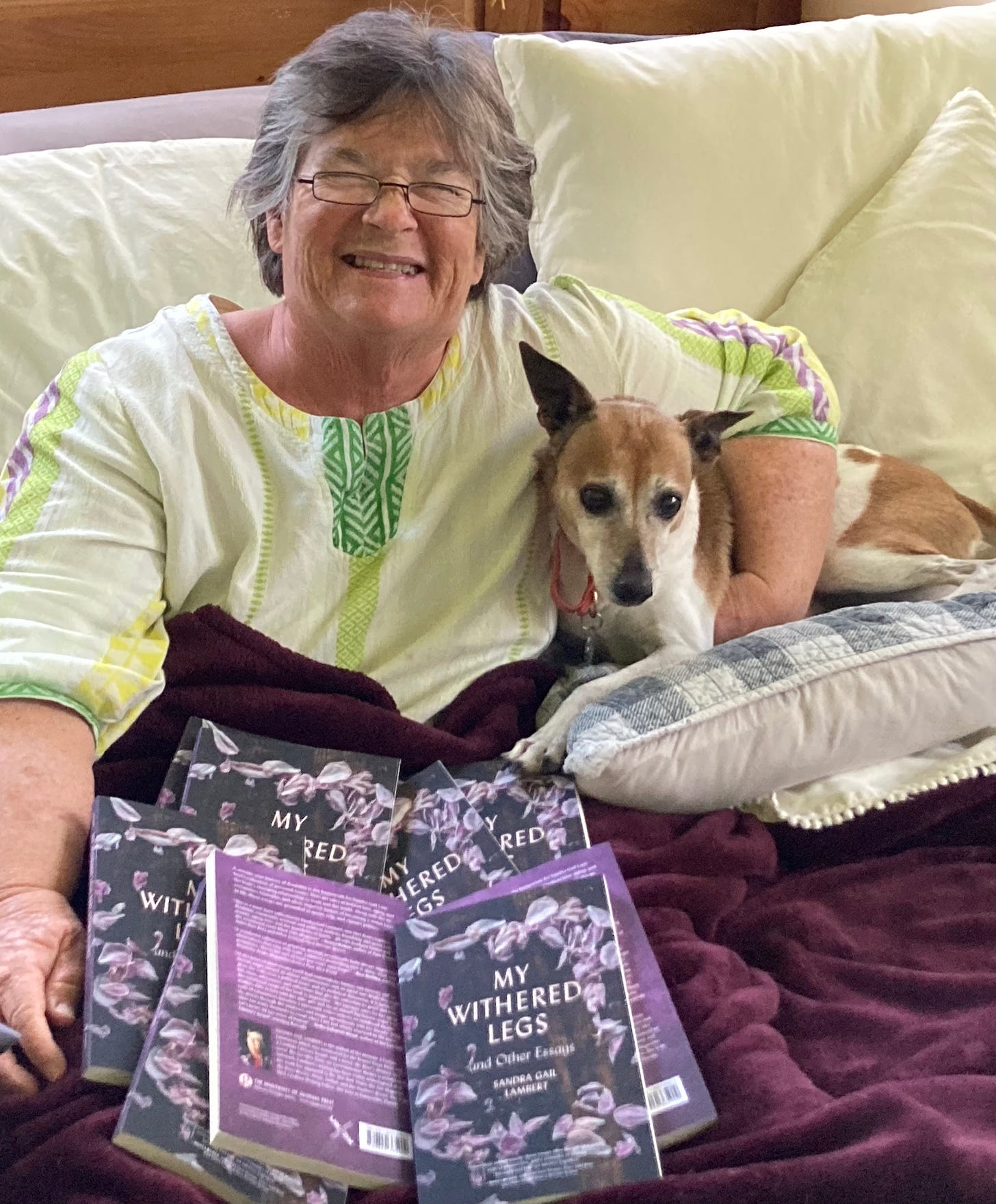 An older white woman in a yellow mu mu sitting propped up in bed with a little brown and white terrier type dog tucked under her arm. Many copies of "My Withered Legs and Other Essays" are spread over her lap. 