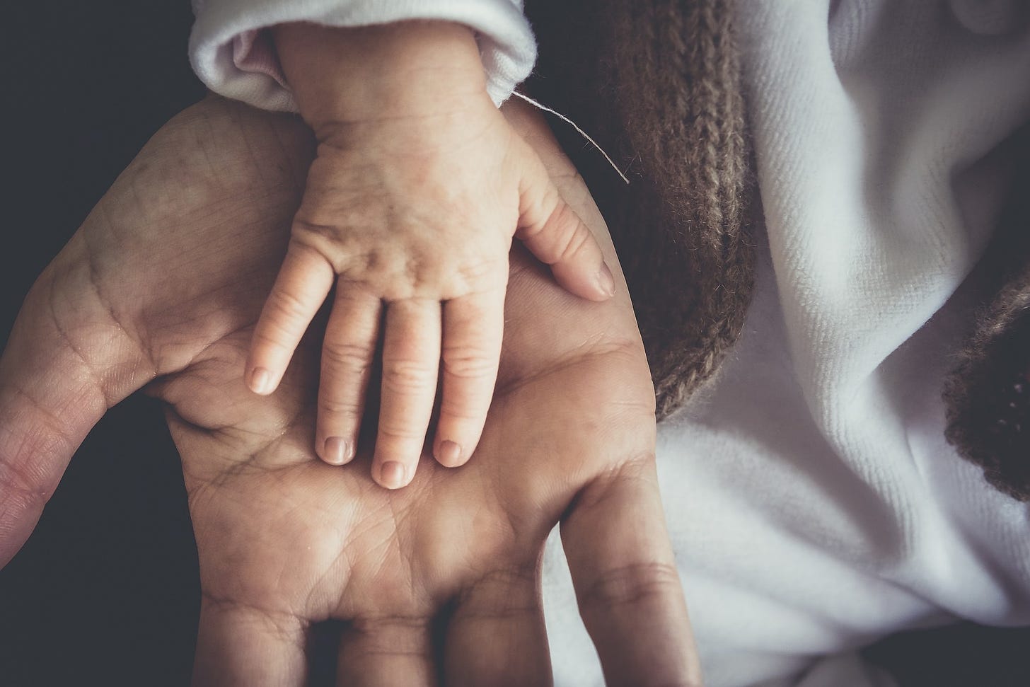 A mão aberta de um adulto branco apoia a mãozinha de uma criança, também branca, abertas palma com palma