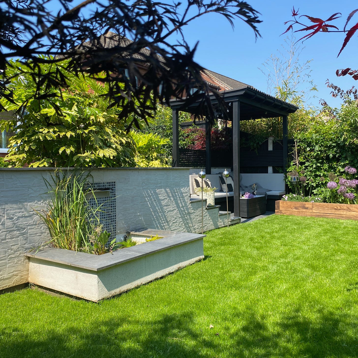 back garden full of greenery and a wooden pergola positioned in the far back right corner and a water feature to the left with pond plants