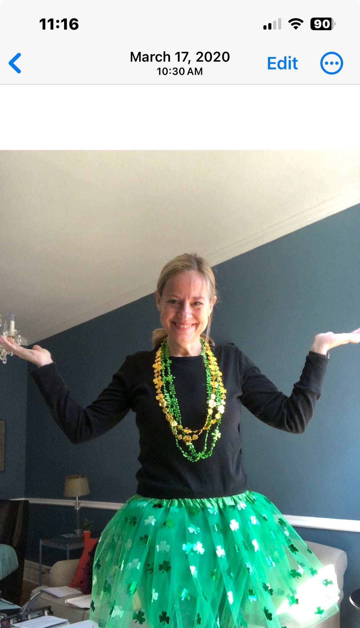 Blonde woman in a black shirt and green skirt. She's wearing green and yellow beads, and there are shamrocks on her skirt for St. Patrick's Day. She is smiling with her hands raised. The walls behind her are blue. 