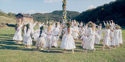 Many women in white dresses and flower crowns raise and lower their arms in a circular fashion around a May pole. 