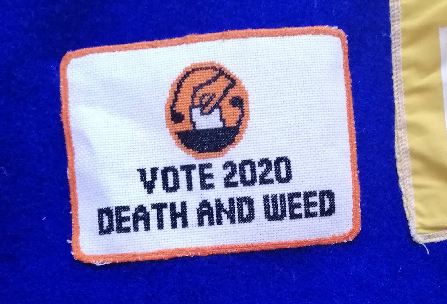 Cross stitch with the Election NZ logo and the text "Vote 2020 Death and Weed" sewn onto a blue wool blanket