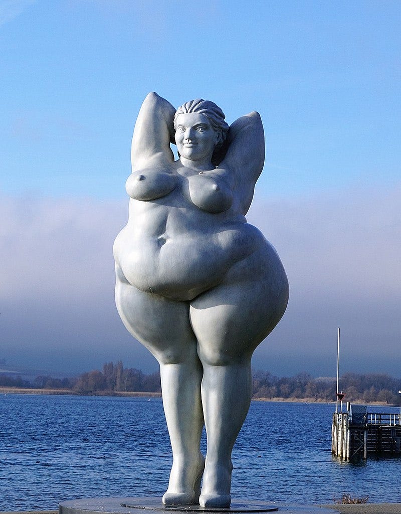 fotografia de ambiente externo. o mar ao fundo e em primeiro plano uma estátua de pedra cinza gigantesca mostrando uma mulher obesa nua, numa posição confiante, com os dois braços jogados para cima e o olhar sereno.