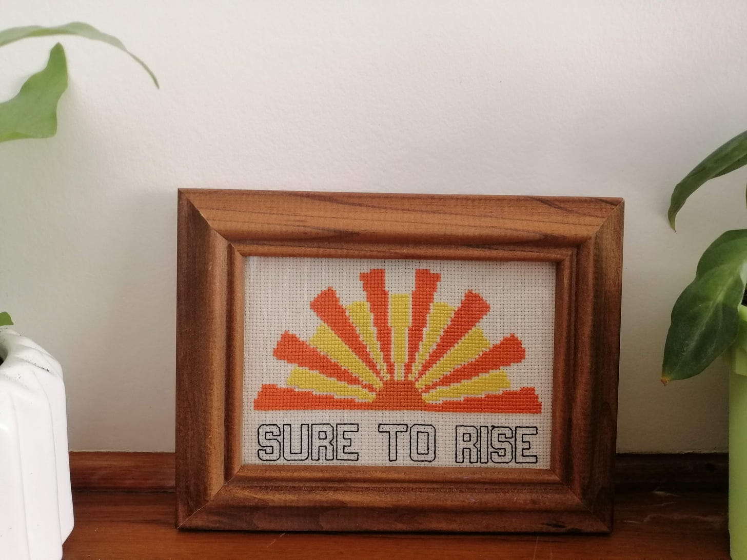 Completed cross stitch in a brown wooden frame. The stitch is the Edmonds rising sun logo with the text "Sure to rise" like the top of the factory.