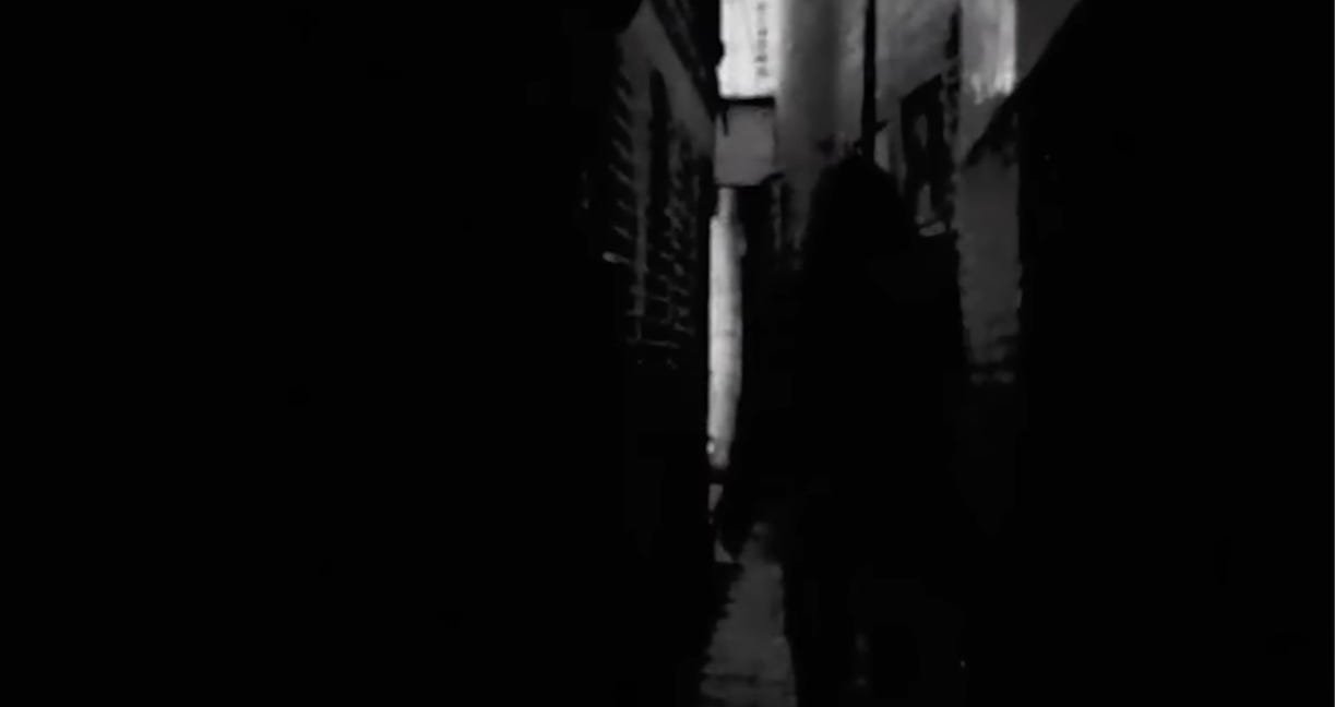 In a dark alley, a shadowy figure in a hood approaches, all in black and white.
