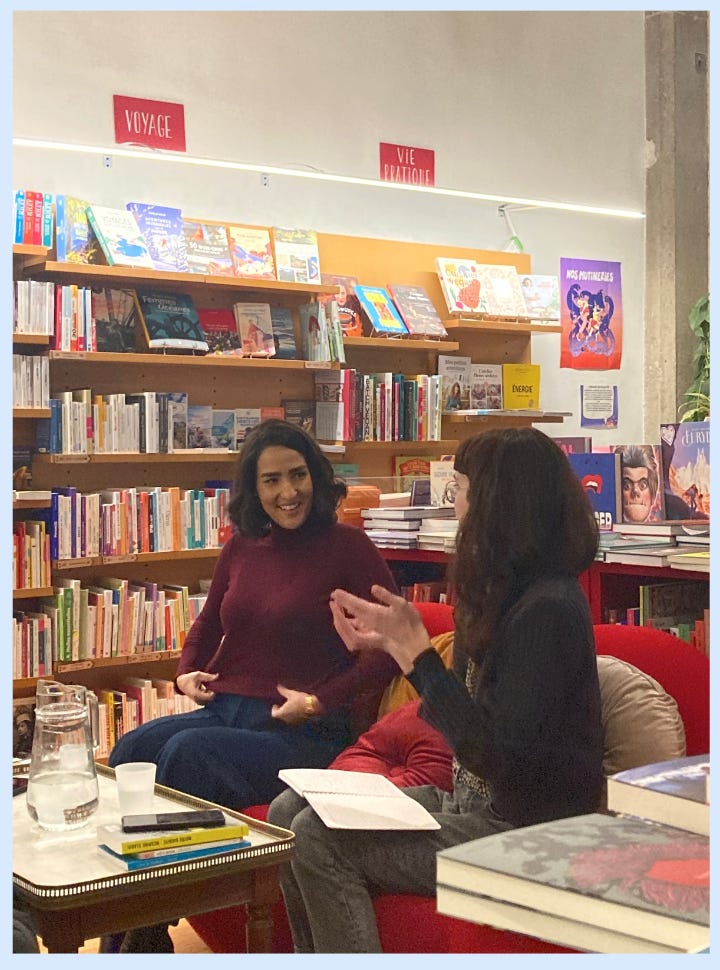 Nesrine Slaoui et la libraire sont installées sur un canapé rouge devant un rayon de livre en train d'échanger avant la rencontre. Devant elles se trouve une carafe d'eau, deux verres et des livres