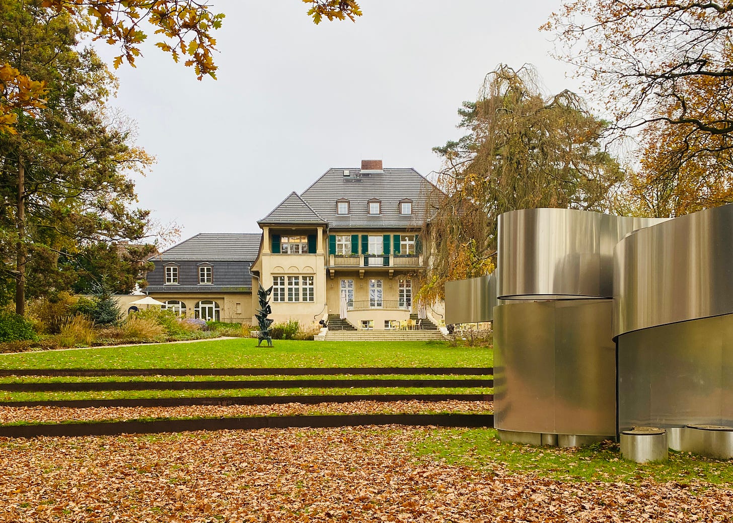  Mansão histórica cercada de gramado e árvores de outono, com uma escultura moderna de metal à direita e folhas caídas cobrindo o chão.