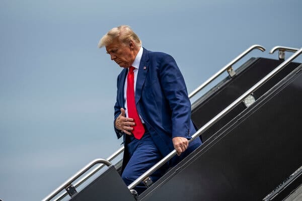 Former President Donald J. Trump, wearing a blue suit and red tie, getting off a plane. 