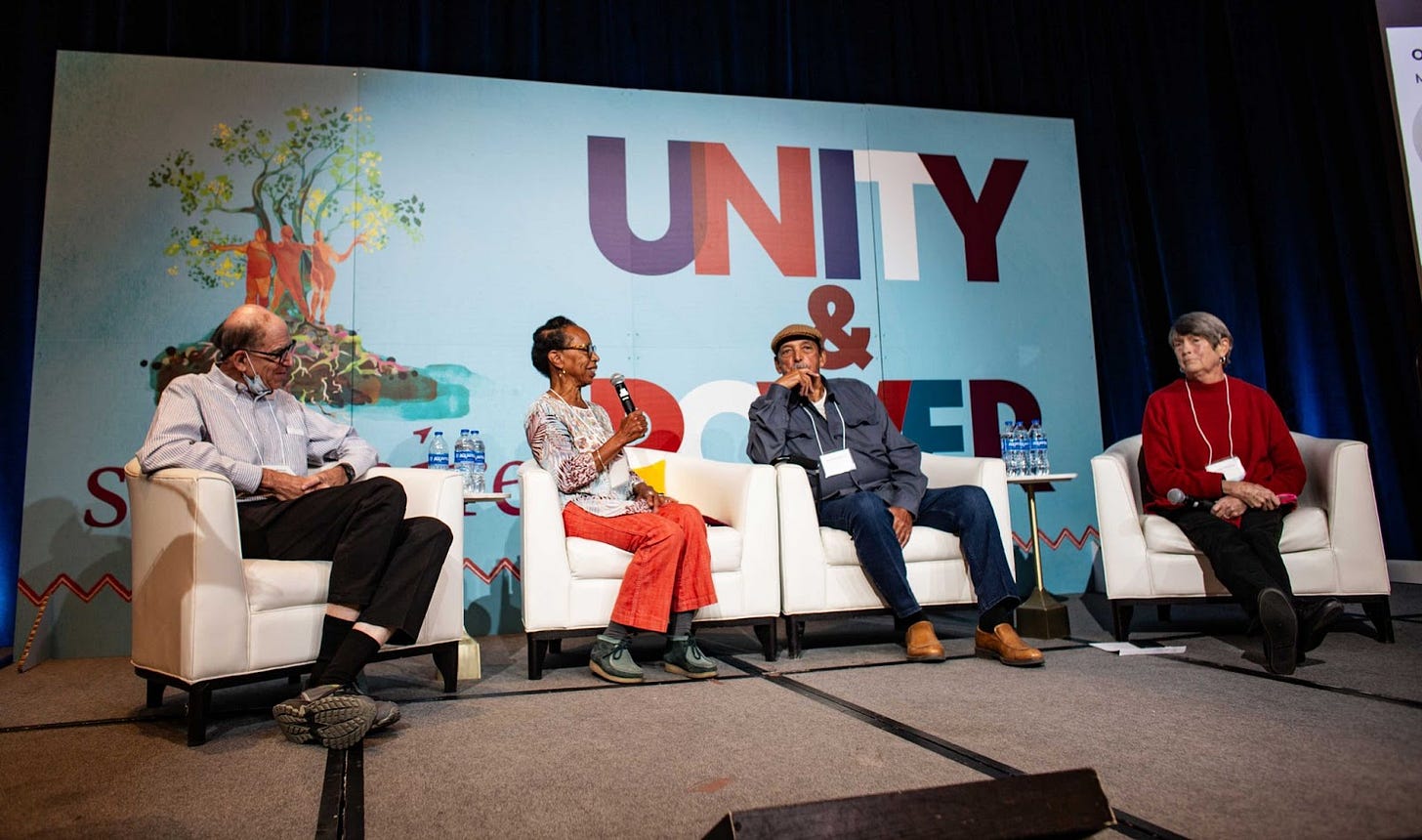 Four people sitting in cushy white chairs on a stage in front of a blue background.