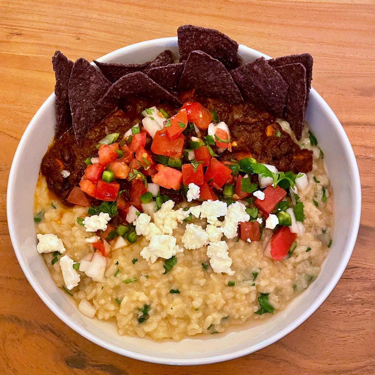 A bowl of herby risotto with a pile of pico de gallo and cheese in the center, and blue corn tortilla chips at the back.