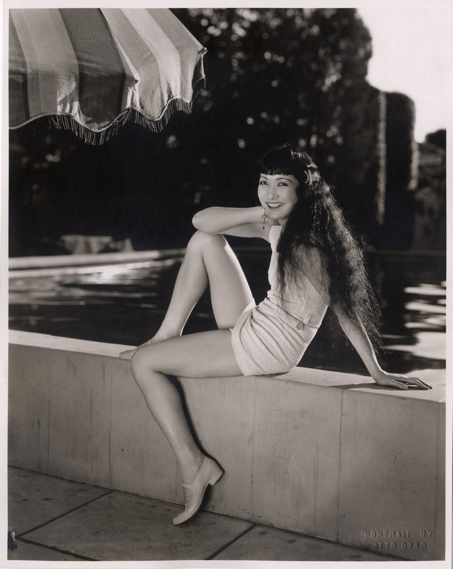 Anna May Wong sits poolside in a bathing suit with her long hair down, smiling