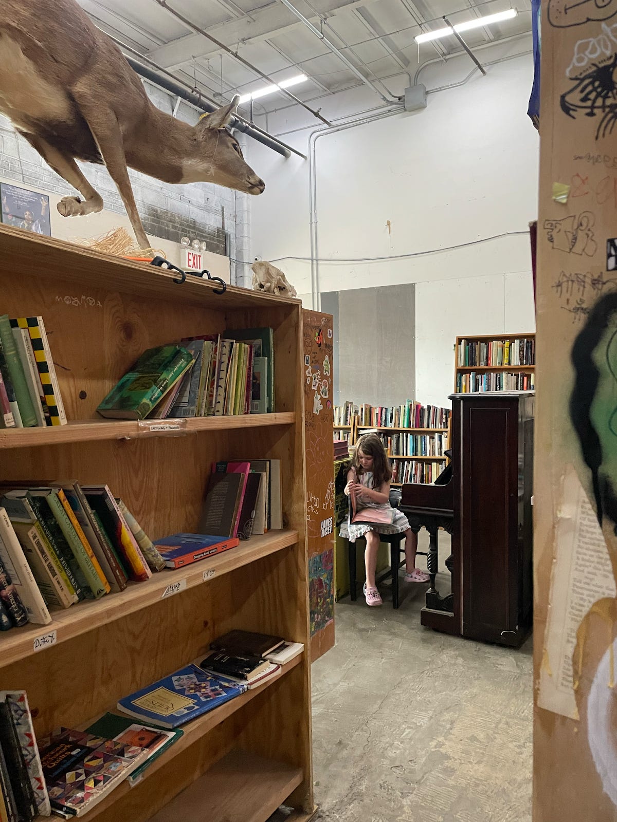 A child flipping through a book sitting on a piano bench. A taxidermy deer is atop a nearby bookshelf.