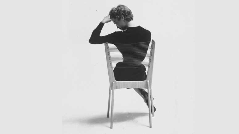 Monochrome 1950s image of the back of a woman in a black dress seated on a chair made of a wood frame and cord strung seat and back.