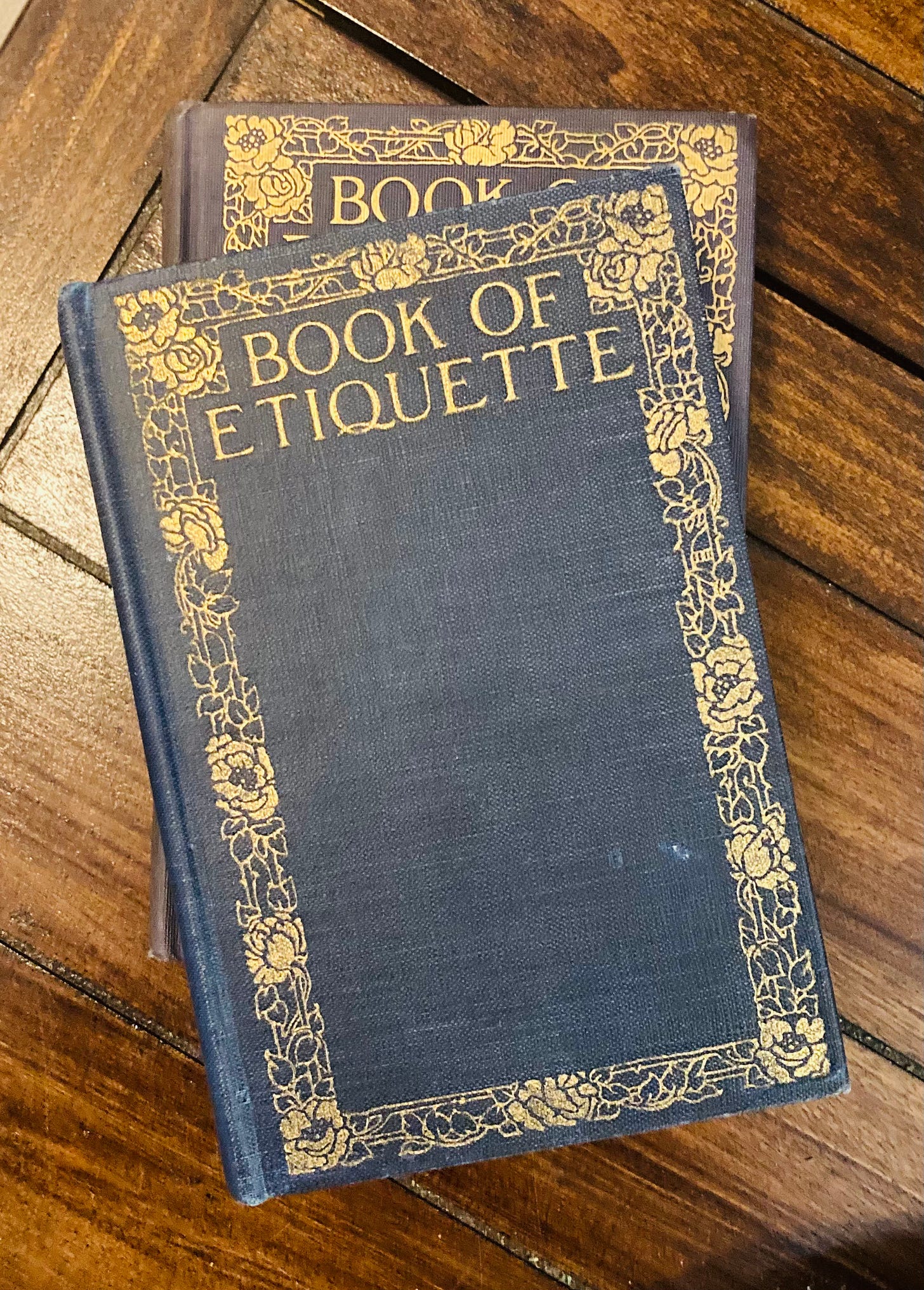 Two cloth bound dark blue books with gold foil writing that reads Book of Etiquette with a rose border detail 