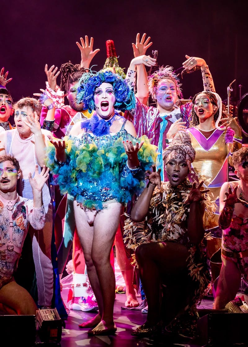 Taylor Mac, a white person in a large, messy, blue and aqua wig and a glittery feathered minidress sings with a wide open mouth, hands outstretched. Around Taylor is a colorful multiracial cast wearing extravagant and varied costumes, signing with arms raised.