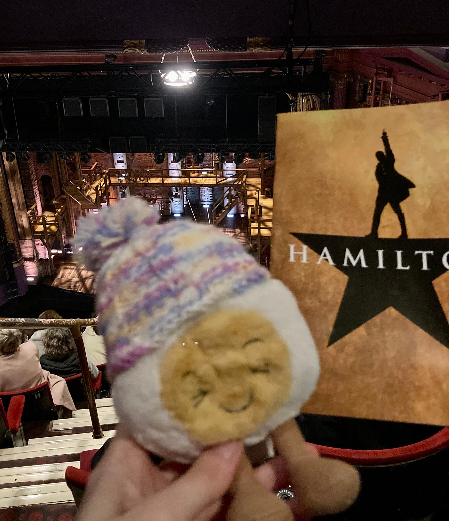 Photo of Dippy, the soft toy egg in a bobble hat, in theatre seats awaiting the musical performance of Hamilton. The stage is set with wooden scaffolding and Dippy is framed by a program displaying the signature Hamilton star silhouette on a gold background.