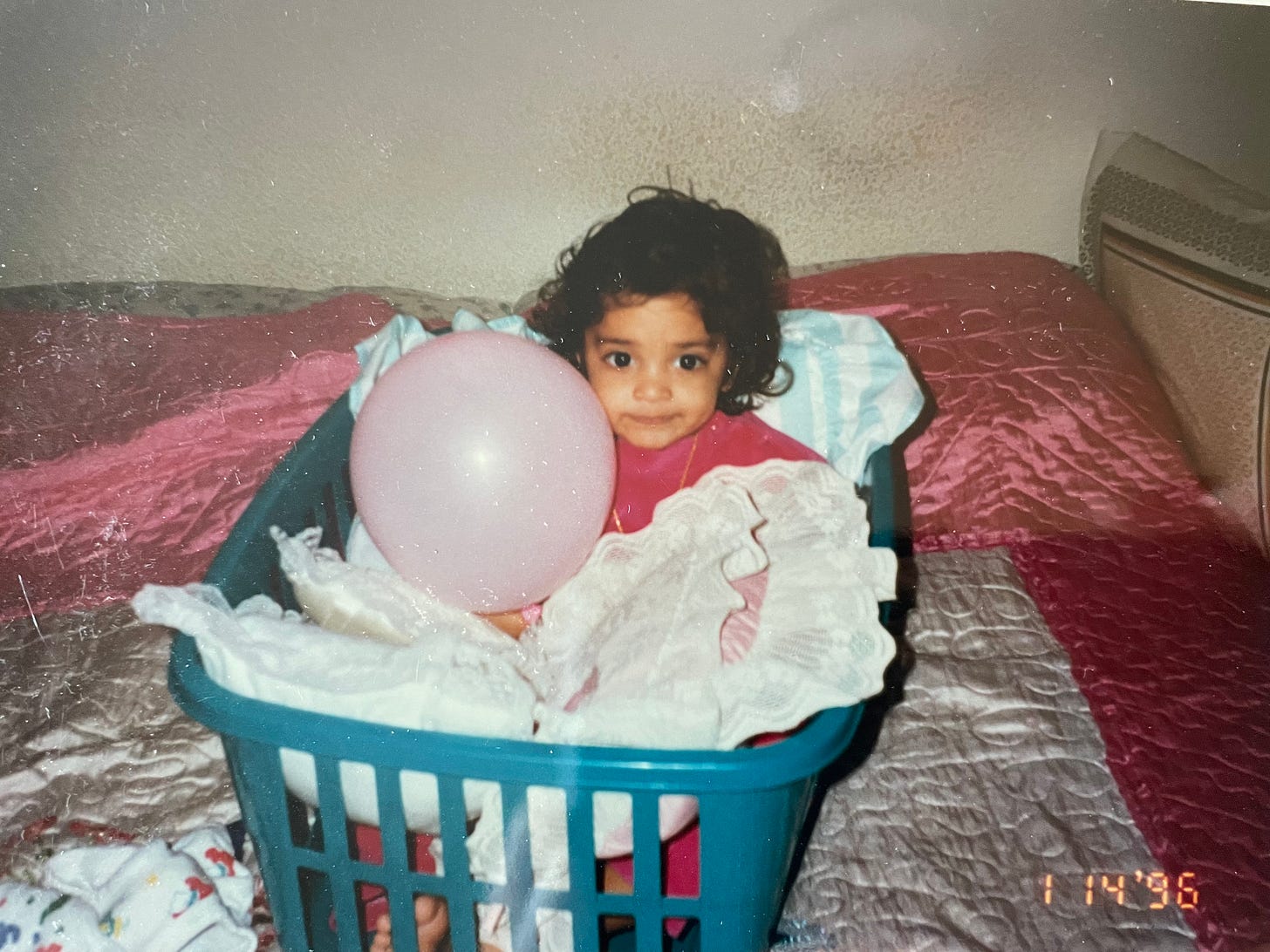 A toddler with curly hair sits inside of a green basket full of pillows. She holds a light pink balloon and is atop a bed with pink and white bedding.