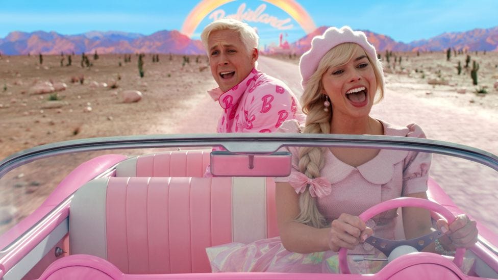 Actors Ryan Gosling and Margot Robbie in a still image taken from a scene in the Barbie movie. They're both in a pink open-top car driving along a road in what looks like a desert. A rainbow can be seen in the background over a mountain range with the words Barbieland underneath it. Margot as Barbie is wearing a pink dress and pink hat with a pink bow in her plaited blonde hair and is sat in the front seat with her hands on the pink steering wheel. Ryan as Ken is sat in the back of the car with his bleach blonde hair and a pink jacket on which is covered in the letter B. Both have their mouths open as if they are singing