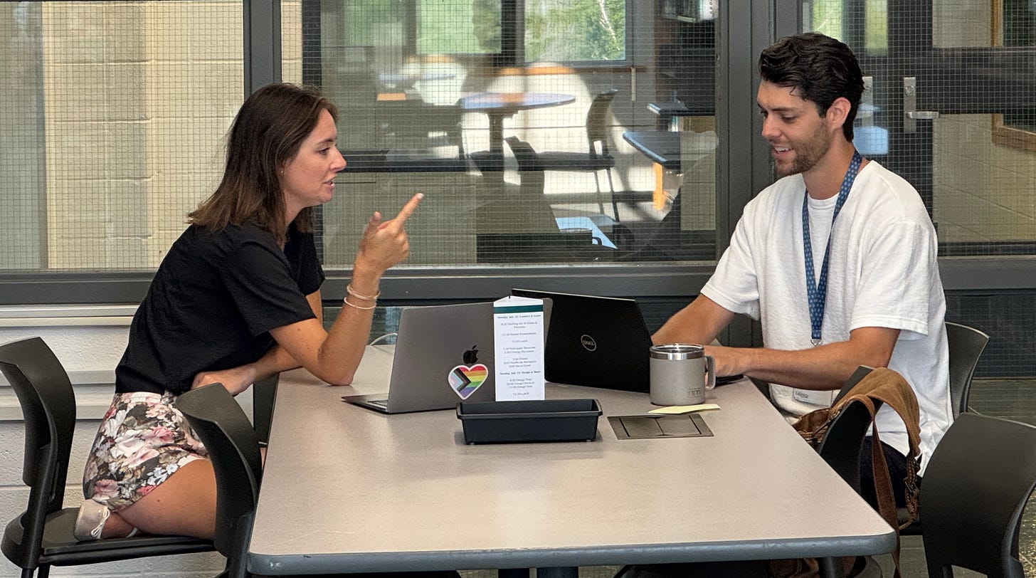 Two people are seated at a table, each with a laptop. The woman on the left is gesturing while speaking, and the man on the right is smiling and looking at his laptop. The table has a small tray with a paper displaying a schedule or list. The background includes large windows with a view of another room.